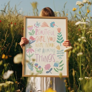 May include: A framed print with the words "BEAUTIFUL GIRL YOU WERE MADE FOR AMAZING THINGS" surrounded by colorful floral illustrations. The frame is a light brown color. The print is held in a field of tall grass and wildflowers.