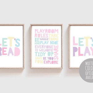 May include: Three framed prints with colourful text. The first print says "Let's Read" in a rainbow of colours. The second print says "Playroom Rules" and lists rules for a playroom, including "Take Turns", "Be Kind", "Play Nice", "Everyone is Welcome", "Tidy Up", "Share", "Have Fun", and "Explore". The third print says "Let's Play" in a rainbow of colours. The prints are on a white background.