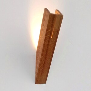 May include: A wooden wall sconce with a warm light shining through the open space in the center. The sconce is made of natural wood with a light brown finish.