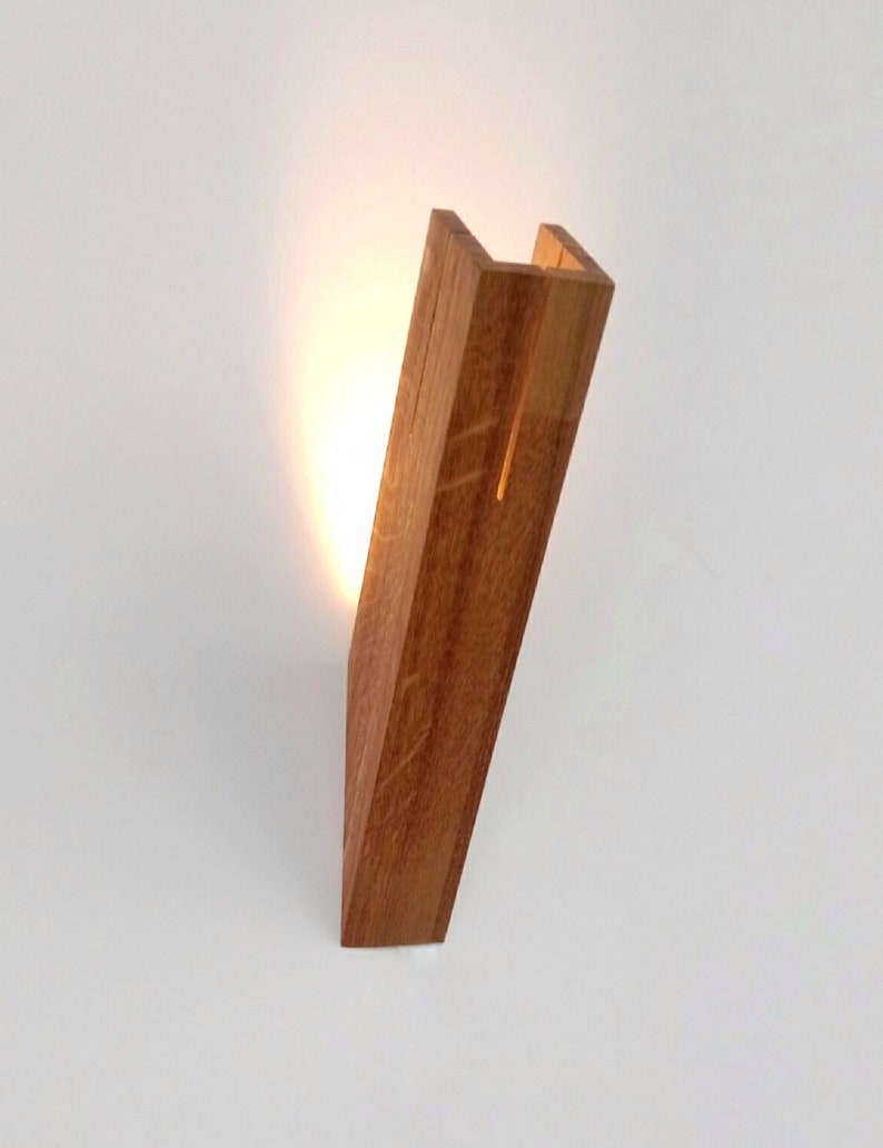 May include: A wooden wall sconce with a warm light shining through the open space in the center. The sconce is made of natural wood with a light brown finish.