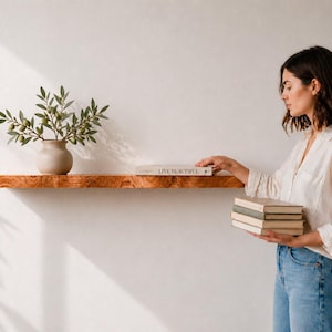 May include: A natural edge wooden floating shelf mounted on a wall, featuring a light brown wood grain. A beige ceramic vase with green olive branches sits on the left, and a book titled LIVE BEAUTIFUL rests on the shelf surface.