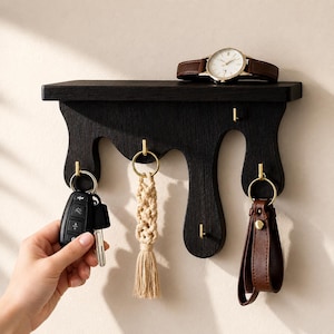 Könnte beinhalten: Ein schwarzes Wandregal aus Holz mit tropfenförmiger Unterkante und fünf goldfarbenen Metallhaken. Auf dem Regal liegt eine Armbanduhr mit braunem Lederarmband, während an den Haken ein Autoschlüssel und ein Makramee-Anhänger hängen.