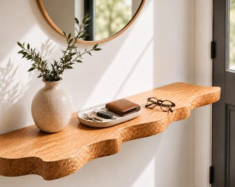 May include: A wall-mounted wooden shelf with a wavy, live-edge front. On the shelf sits a round, textured beige ceramic vase containing green leafy branches, an oval ceramic tray holding a brown leather wallet and keys, and tortoise-shell glasses.