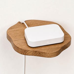 May include: A white rectangular electronic device with a small indicator light sits on a natural-edged wooden wall shelf. A thin white power cord extends from the back of the device, running down the wall behind the wooden shelf surface.