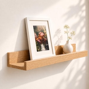 May include: A light oak wood floating wall shelf featuring a raised front edge. A white picture frame containing a floral print and a small white ceramic vase holding delicate white flowers are positioned on the shelf surface.