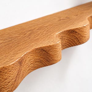 May include: A light brown wooden shelf featuring a distinct scalloped edge design. The surface displays a visible natural wood grain pattern with a smooth finish, highlighting the texture and warm tones of the timber material.