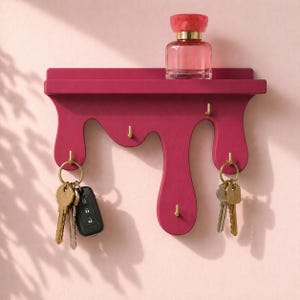 May include: A magenta wall-mounted shelf with a dripping paint design featuring five gold-toned metal hooks. A clear glass perfume bottle with a pink cap sits on the top shelf, while two sets of metal keys hang from the outer hooks.