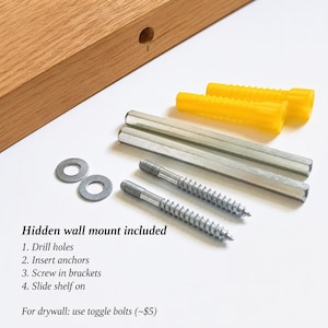 May include: A light brown wooden shelf with a pre-drilled hole, two yellow plastic wall anchors, two silver metal cylindrical brackets, two silver metal screws, and two silver metal washers. Text reads: Hidden wall mount included.