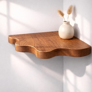 May include: A wall-mounted corner shelf made of medium-toned wood with a wavy, cloud-like front edge. A small, round, off-white ceramic vase containing two dried, fluffy, light brown botanical stems sits on the surface of the wooden shelf.