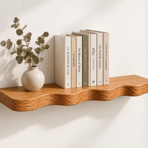 May include: A wall-mounted wooden shelf with a wavy front edge, holding a small round ceramic vase with green eucalyptus stems and a row of seven books with neutral-colored spines titled The Kinfolk Home, At Home, The Beauty of Home, Live Beautifully, Still, The Kinfolk Garden, and A Life More Considered.