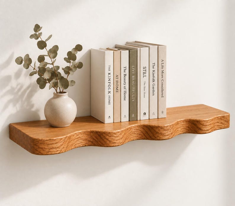 May include: A wall-mounted wooden shelf with a wavy front edge, holding a small round ceramic vase with green eucalyptus stems and a row of seven books with neutral-colored spines titled The Kinfolk Home, At Home, The Beauty of Home, Live Beautifully, Still, The Kinfolk Garden, and A Life More Considered.