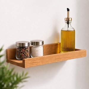Könnte beinhalten: Ein rechteckiges Wandregal aus hellbraunem Holz mit einem durchsichtigen Ölspender aus Glas mit goldenem Metallausgießer, einem kleinen Glas mit bunten Pfefferkörnern und einem kleinen Glas mit weißem Salz, alle mit silberfarbenen Deckeln.