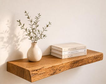 May include: A natural edge wooden floating shelf holding a cream-colored ceramic vase with green leafy branches and a stack of three books with the titles This Is Home, Live Beautiful, and be well printed on the spines in a neutral color palette.