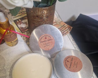May include: Three silver tins of rosehip salve, one open, revealing a light beige balm. The tins have brown labels with the text "Rosehip Remedies Rosehip Salve". A bottle of rosehip infused oil and a vase of white roses are in the background.