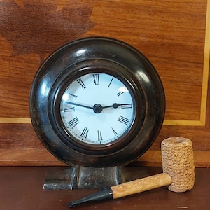 May include: A dark brown wooden clock with Roman numeral markings on a white face. The clock is on a wooden stand and a brown and black corncob pipe is in the foreground.