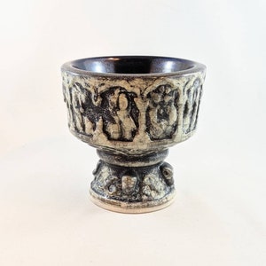 Puede incluir: Un cuenco de cerámica con un esmalte blanco y negro. El cuenco tiene un borde elevado con un diseño de figuras y arcos. El cuenco se asienta sobre un pedestal con un diseño similar.