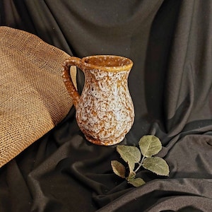 Puede incluir: Una taza de cerámica marrón con un patrón moteado blanco. La taza tiene un asa y una abertura ancha. Un saco de arpillera y una ramita de hojas verdes están en el fondo. La taza mide aproximadamente 13 cm de alto.