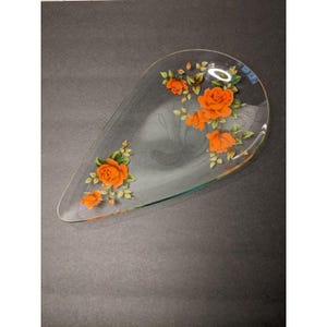 May include: Clear glass serving dish in a teardrop shape, adorned with orange rose floral designs and green leaves. The dish has a smooth surface and a slightly raised edge. The floral pattern is hand-painted, adding a touch of elegance to the piece.