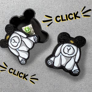 May include: Black and white bear-shaped container with a matching lid, and a separate bear-shaped item. The container is open, revealing a small green item inside. The word "CLICK" is written in yellow.