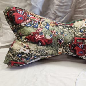 May include: A decorative pillow with a patriotic theme. The pillow features a print of red trucks, American flags, and white flowers on a green background. It has a bow tie shape and a strap with a metal clip.