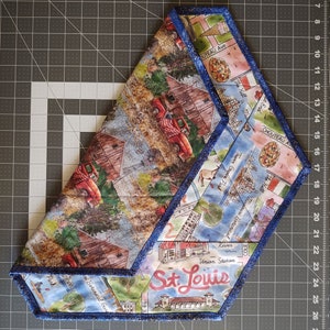 Può includere: Un tessuto blu e bianco con un design a patchwork che presenta un camion rosso, un fienile e una città con il testo "St. Louis". Il tessuto ha un bordo blu.