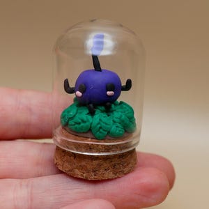 Op de afbeelding: Een miniatuur paars wezen met zwarte antennes en armen zit bovenop groene bladeren in een heldere glazen koepel. De koepel rust op een kurken voet. Het kunstwerk wordt in een hand gehouden.