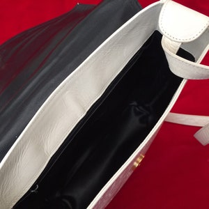 Puede incluir: Bolso de hombro de cuero negro y blanco abierto. El bolso tiene una solapa y correa blancas, un interior negro y un cierre dorado. El bolso est&aacute; sobre un fondo rojo.