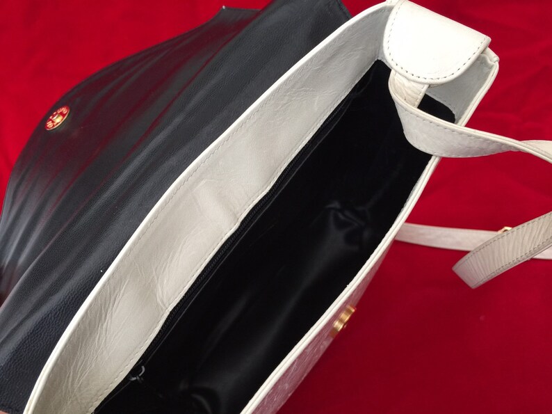 Puede incluir: Bolso de hombro de cuero negro y blanco abierto. El bolso tiene una solapa y correa blancas, un interior negro y un cierre dorado. El bolso est&aacute; sobre un fondo rojo.