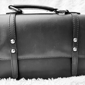 Könnte beinhalten: Schwarze Umhängetasche mit Tragegriff und verstellbarem Schulterriemen. Die Tasche hat silberfarbene Nieten und dekorative Riemen auf der Vorderseite. Der Klappenverschluss sichert das Hauptfach. Die Tasche ist aus glattem, lederähnlichem Material.