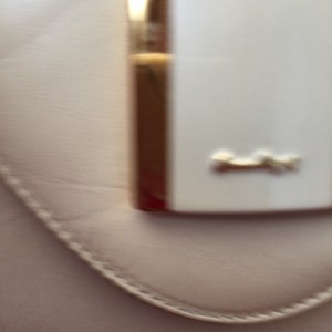 Puede incluir: Primer plano de un bolso rosa claro con un cierre rectangular blanco y herrajes dorados. El cierre tiene un peque&ntilde;o emblema dorado. El bolso tiene un borde cosido.