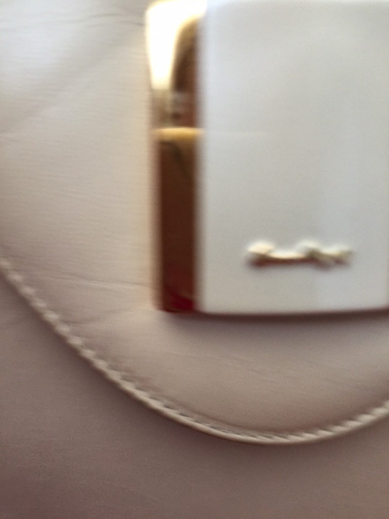 Puede incluir: Primer plano de un bolso rosa claro con un cierre rectangular blanco y herrajes dorados. El cierre tiene un peque&ntilde;o emblema dorado. El bolso tiene un borde cosido.