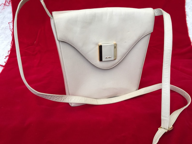 Puede incluir: Bolso bandolera de cuero color crema con forma trapezoidal y cierre de solapa. El bolso presenta un cierre rectangular dorado y una correa larga y fina para el hombro. El bolso est&aacute; sobre un fondo rojo.