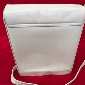 Puede incluir: Bolso bandolera rectangular blanco con una correa larga y fina. El bolso tiene un cierre de solapa y una superficie texturizada. El fondo es de color rojo liso.