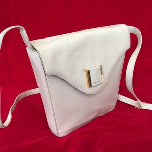 Puede incluir: Un bolso bandolera de cuero blanco con cierre de solapa y una correa larga y ajustable. El bolso presenta un cierre rectangular dorado en la parte delantera. El bolso est&aacute; sobre un fondo rojo.