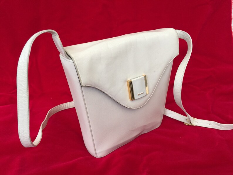 Puede incluir: Un bolso bandolera de cuero blanco con cierre de solapa y una correa larga y ajustable. El bolso presenta un cierre rectangular dorado en la parte delantera. El bolso est&aacute; sobre un fondo rojo.