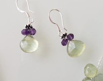 May include: A pair of silver earrings with green and purple gemstone drops. The earrings feature a teardrop-shaped green stone with a faceted cut and a cluster of three small purple stones.