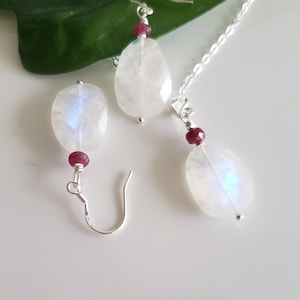 May include: A silver necklace and earrings set featuring a white moonstone pendant and matching earrings with a small red ruby accent.