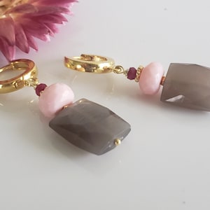 Peut inclure: Une paire de boucles d'oreilles créoles dorées avec un pendentif en pierre rose et grise. Les pierres sont facettées et ont une finition chatoyante.