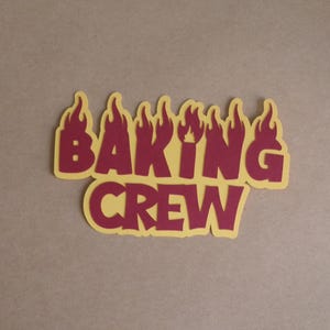 May include: A two-tone paper cutout with the words "BAKING CREW" in a bold, stylized font. The letters are a deep red, outlined with a yellow flame design, set against a neutral background.