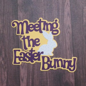 May include: A layered paper cutout with the text "Meeting the Easter Bunny." The text is purple with a yellow background. A white bunny with purple polka dots is in the center of the design. The background is a dark wood grain.