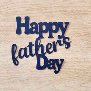 May include: Navy blue cutout that reads "Happy father's Day" in a cursive font. The cutout is placed on a light wood grain surface. The letters are connected and have a rounded, playful style.