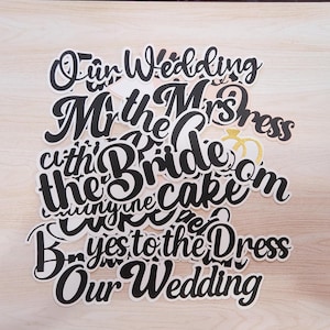 May include: A collection of wedding-themed cake toppers and decorations. The toppers feature phrases like "Our Wedding," "Mr & Mrs," and "Yes to the Dress" in a black script font with white outlines. Some toppers include gold glitter accents.