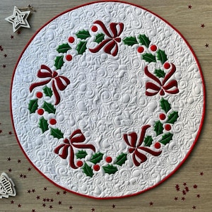 May include: A round, white quilted Christmas tree skirt with a red border. The skirt features a holly and bow design in red and green. The background has a swirling pattern.
