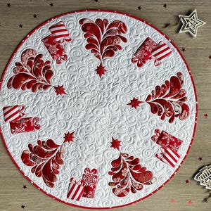 May include: A circular white fabric mat featuring a quilted swirl pattern and a red border. The design includes red and white patterned Christmas tree shapes and gift box motifs arranged in a radial pattern around the center of the mat.