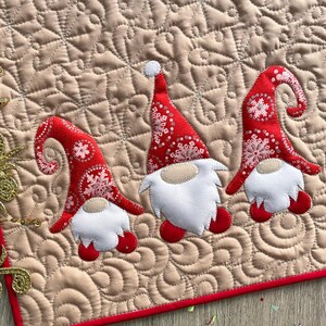 May include: A square quilted fabric mat featuring a beige background with a swirling stitched pattern and a red border. Three gnome appliqués are centered on the mat, each wearing a red hat with a white snowflake print and a white beard.