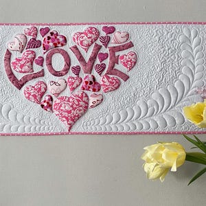 Pode incluir: Uma tapeçaria de parede acolchoada branca com uma borda rosa. A palavra "LOVE" é escrita em letras rosa dentro de uma forma de coração, rodeada por várias peças de tecido em forma de coração rosa. O fundo apresenta um design de penas acolchoadas.