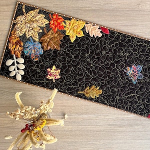 Op de afbeelding: Een rechthoekige, zwart gewatteerde tafelloper met geappliqueerde herfstbladeren in verschillende kleuren. De bladeren bevatten tinten goud, oranje, rood en groen. De loper is afgezet met een gestippelde stof en heeft ingewikkelde stiksels.