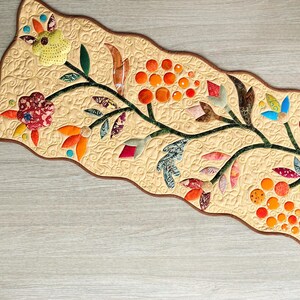 May include: A rectangular table runner with a wavy brown border and a cream-colored background featuring intricate swirl quilting. The runner displays a central vine pattern with colorful fabric appliqué flowers, leaves, and orange circular clusters.