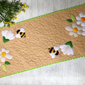 May include: A beige quilted table runner with a green polka dot border. The runner is decorated with white daisies, green leaves, and cartoon bees. The daisies have yellow centers. The runner is approximately 30 inches long.