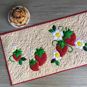 May include: A quilted table runner with a cream background and a red dotted border. The runner is decorated with appliqued strawberries and white flowers. A cinnamon roll sits on a white plate in the upper left corner.